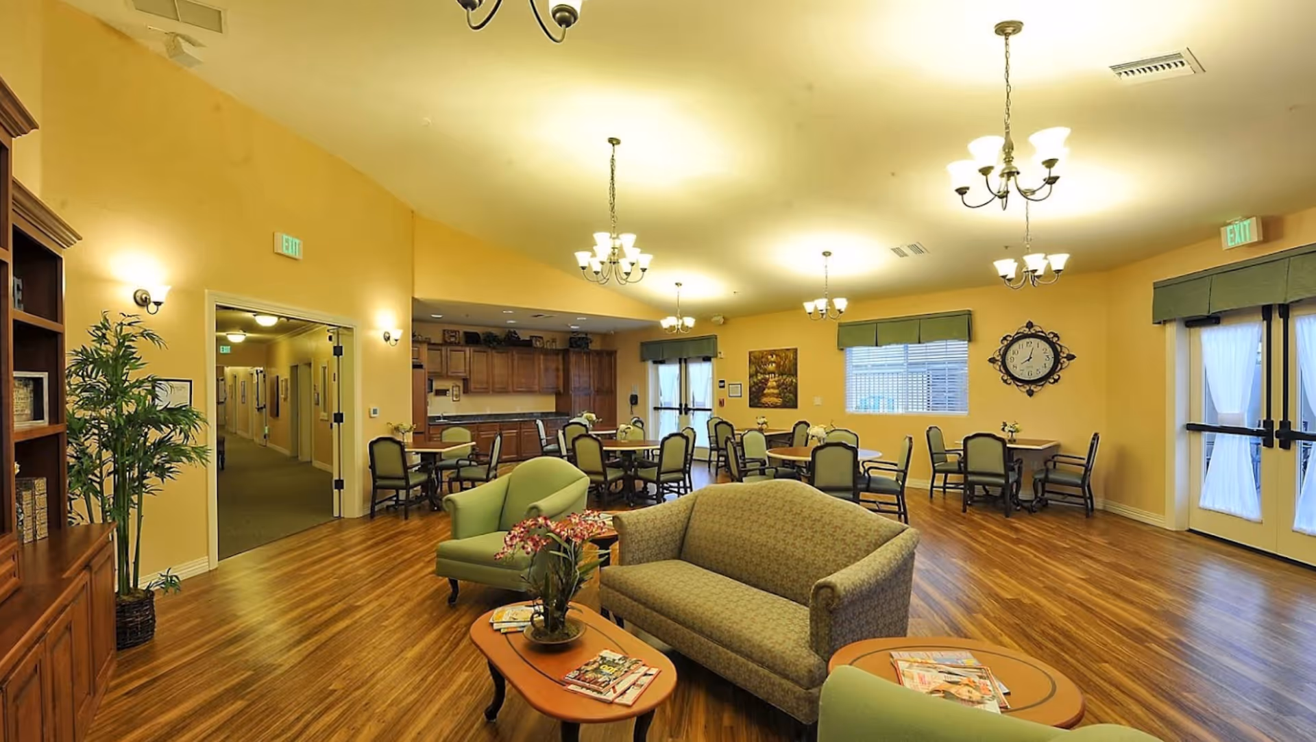 A spacious and well-lit common area with wooden flooring, yellow walls, and multiple chandeliers. The room features several round tables with chairs, a green armchair, a patterned sofa, and a coffee table with magazines and a flower arrangement. There is a large clock on the wall, windows with green valances, and double doors with sheer white curtains. A hallway is visible through an open doorway on the left side.