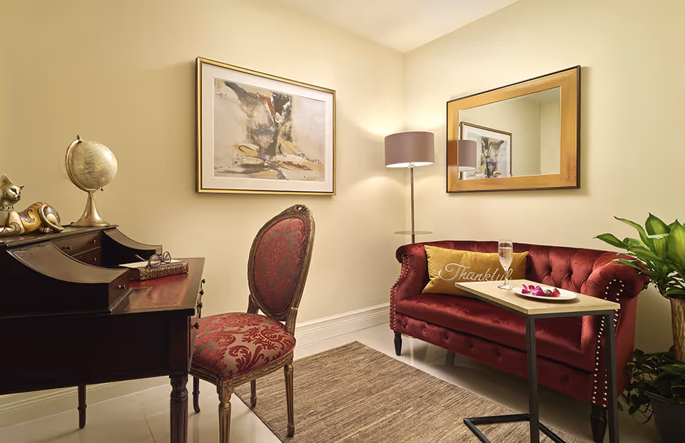 A cozy sitting area with a vintage wooden desk and an ornate red upholstered chair on the left. On the desk are a decorative globe, a cat figurine, and a pair of glasses resting on a book. To the right, there is a plush red velvet loveseat with a gold pillow that says 'Thankful,' a small table holding a glass of water and a plate with flowers, a tall floor lamp, a large gold-framed mirror on the wall, and a green potted plant. The walls are painted a soft beige color and a framed abstract painting hangs above the desk.