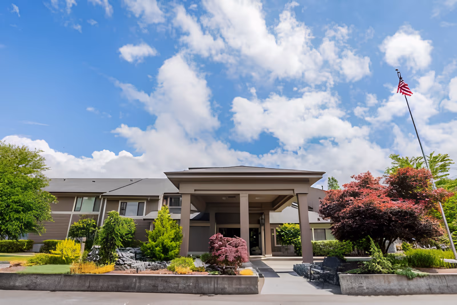 Front exterior view of Village Concepts of Sedro-Woolley - Country Meadow Village building with a covered entrance, surrounded by landscaped greenery and trees under a partly cloudy blue sky. An American flag is visible on a tall flagpole to the right.