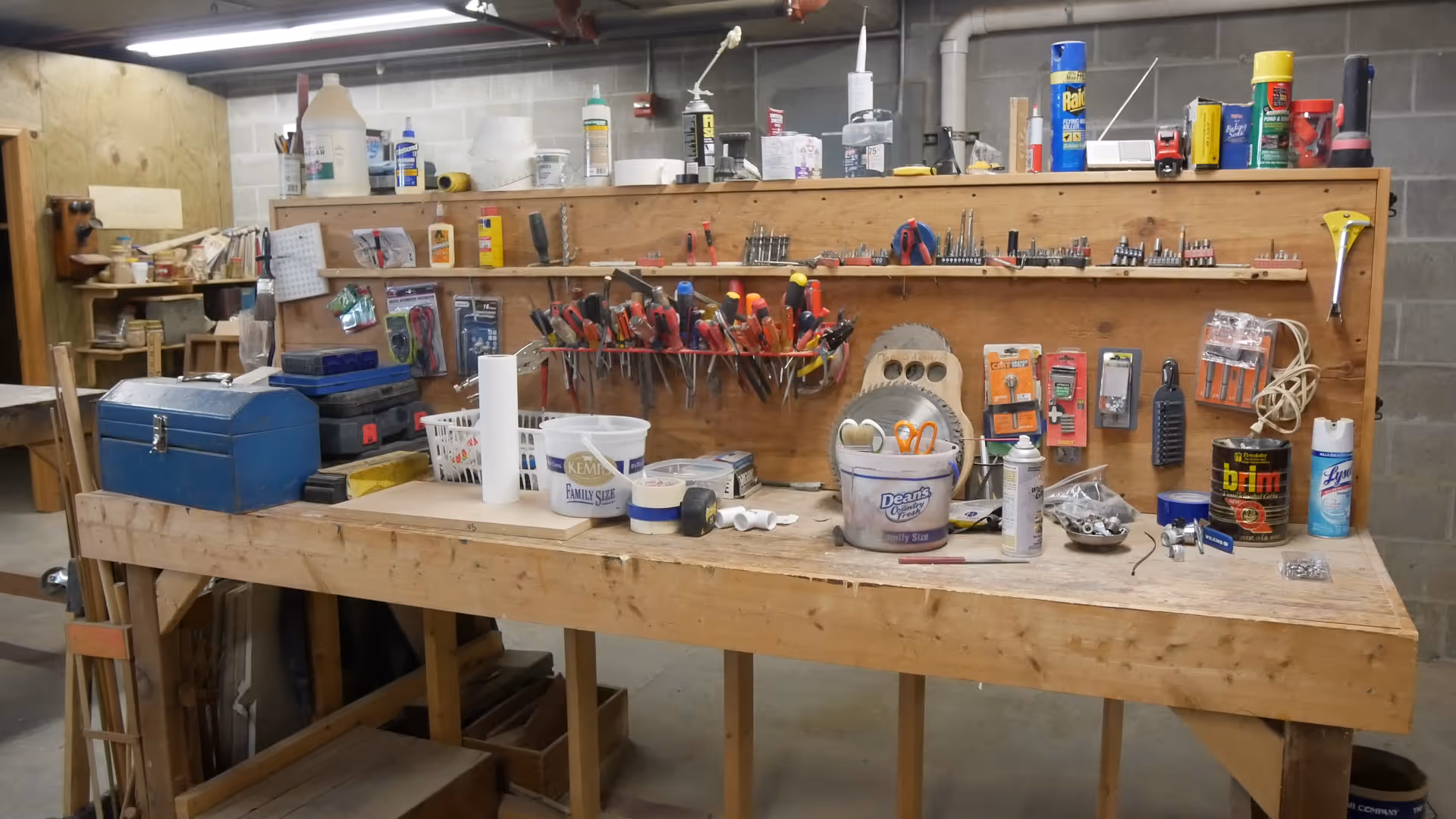 A workshop area with a wooden workbench covered with various tools and supplies. The back wall has a pegboard holding multiple screwdrivers, pliers, and other hand tools. The workbench surface holds containers, cans, a tape measure, scissors, and other small items. The background shows a concrete block wall and additional shelves with more tools and materials.