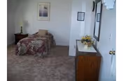 A simple bedroom with a single bed covered in a floral bedspread, a wooden nightstand with a lamp, a framed picture on the wall, and a wooden dresser with a vase of flowers on top.