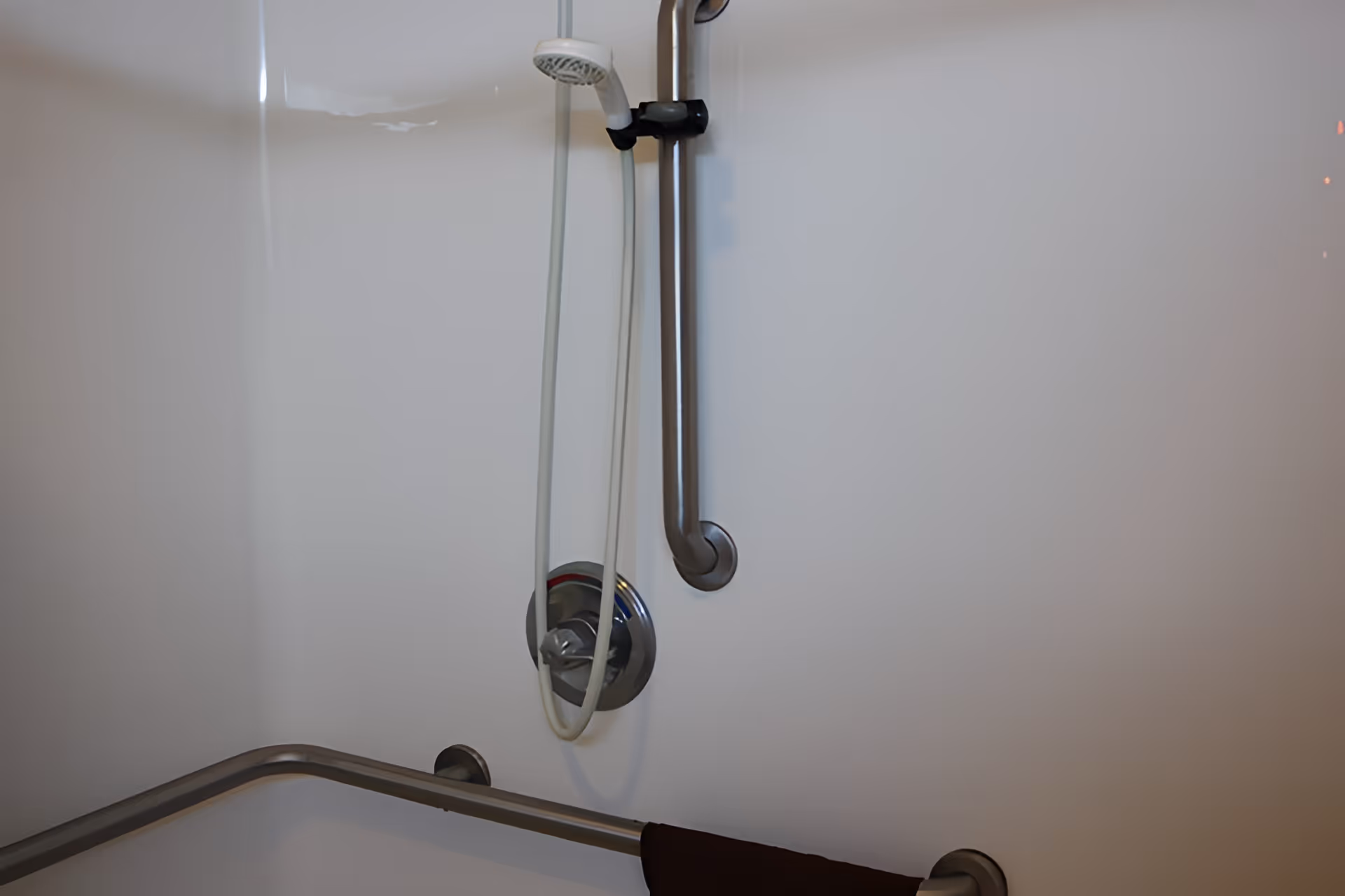 Close-up view of a shower area with a handheld showerhead attached to a vertical sliding bar, a control knob, and two horizontal grab bars installed on the wall for support.
