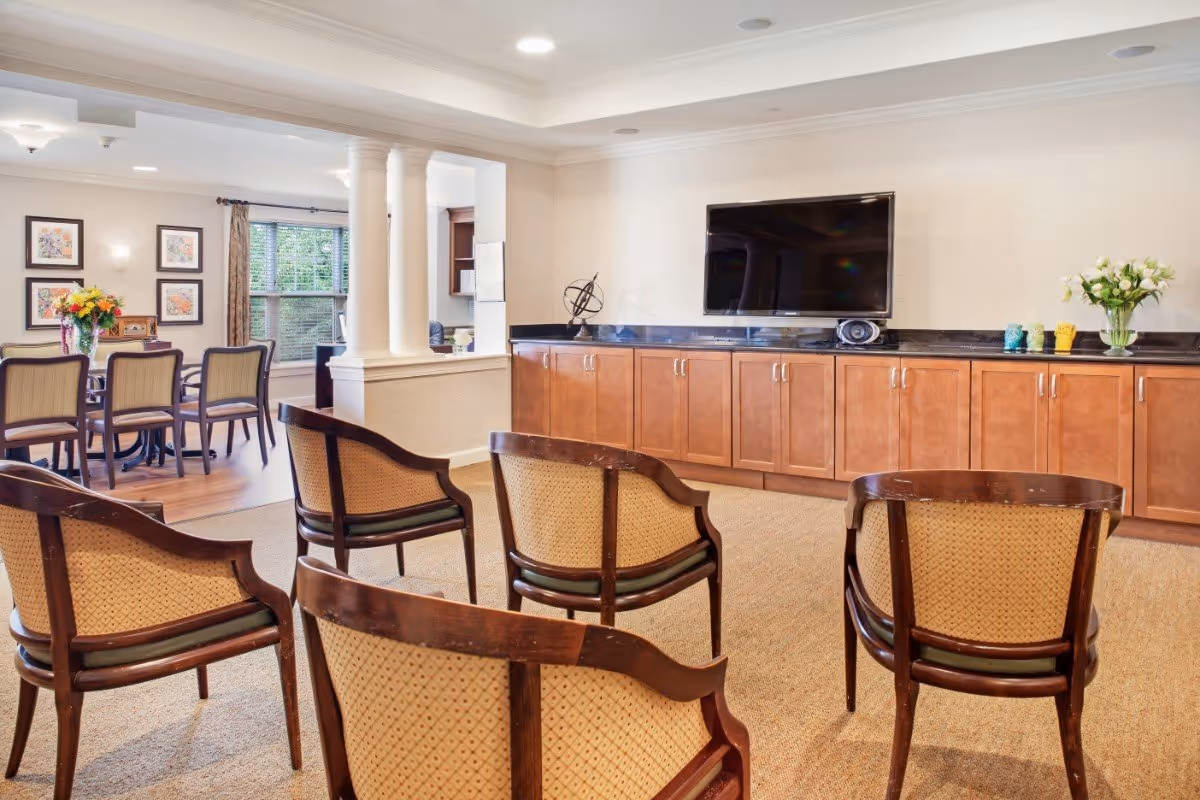 A bright and spacious common area with several wooden chairs arranged facing a large flat-screen TV mounted on a wall above wooden cabinets. To the left, there is a dining area with a table and chairs, decorated with a vase of flowers and framed artwork on the wall. The room has large windows with curtains allowing natural light to enter.