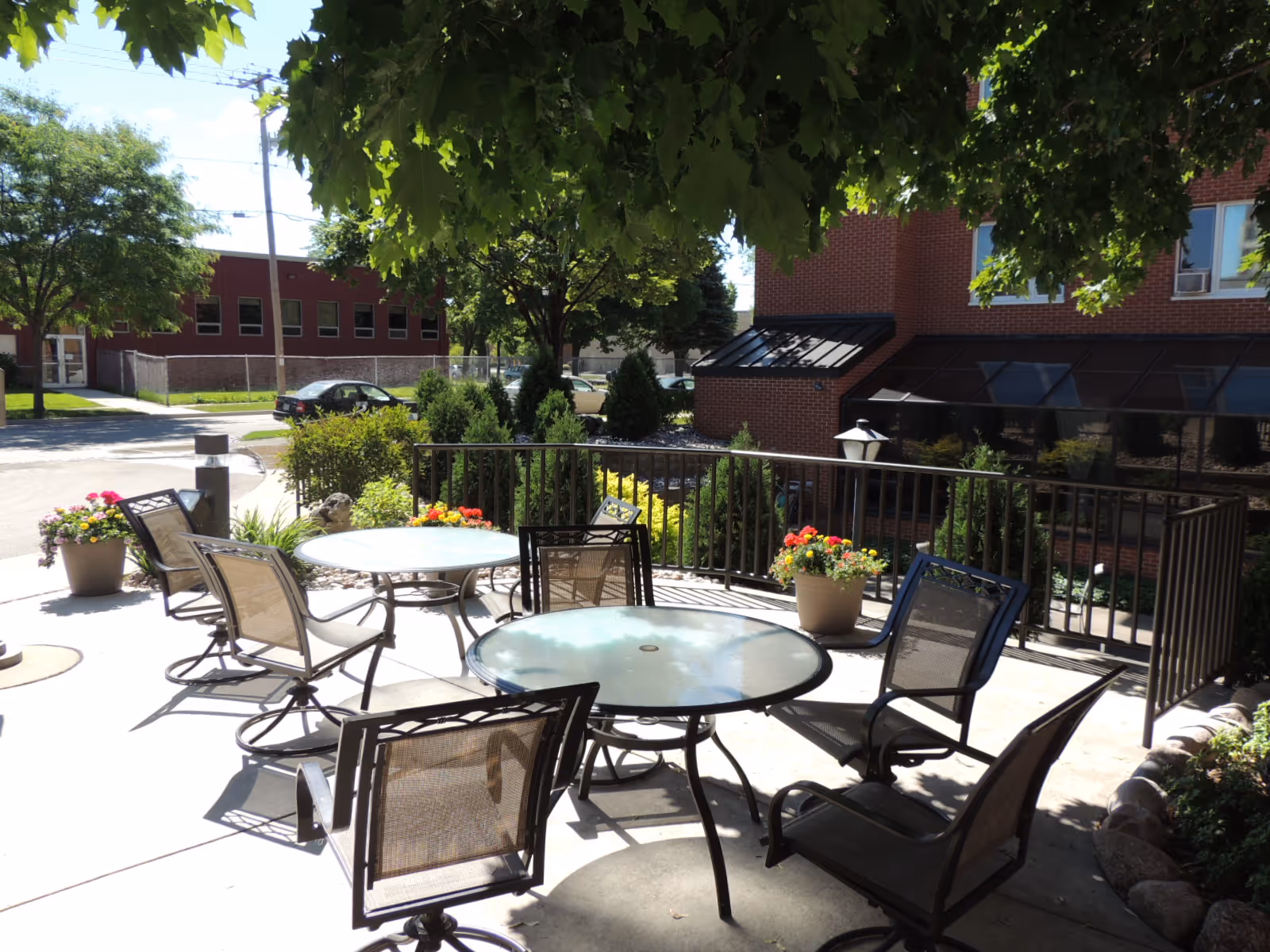 Outdoor patio with round glass tables, metal chairs, potted flowers and trees beside a brick building.