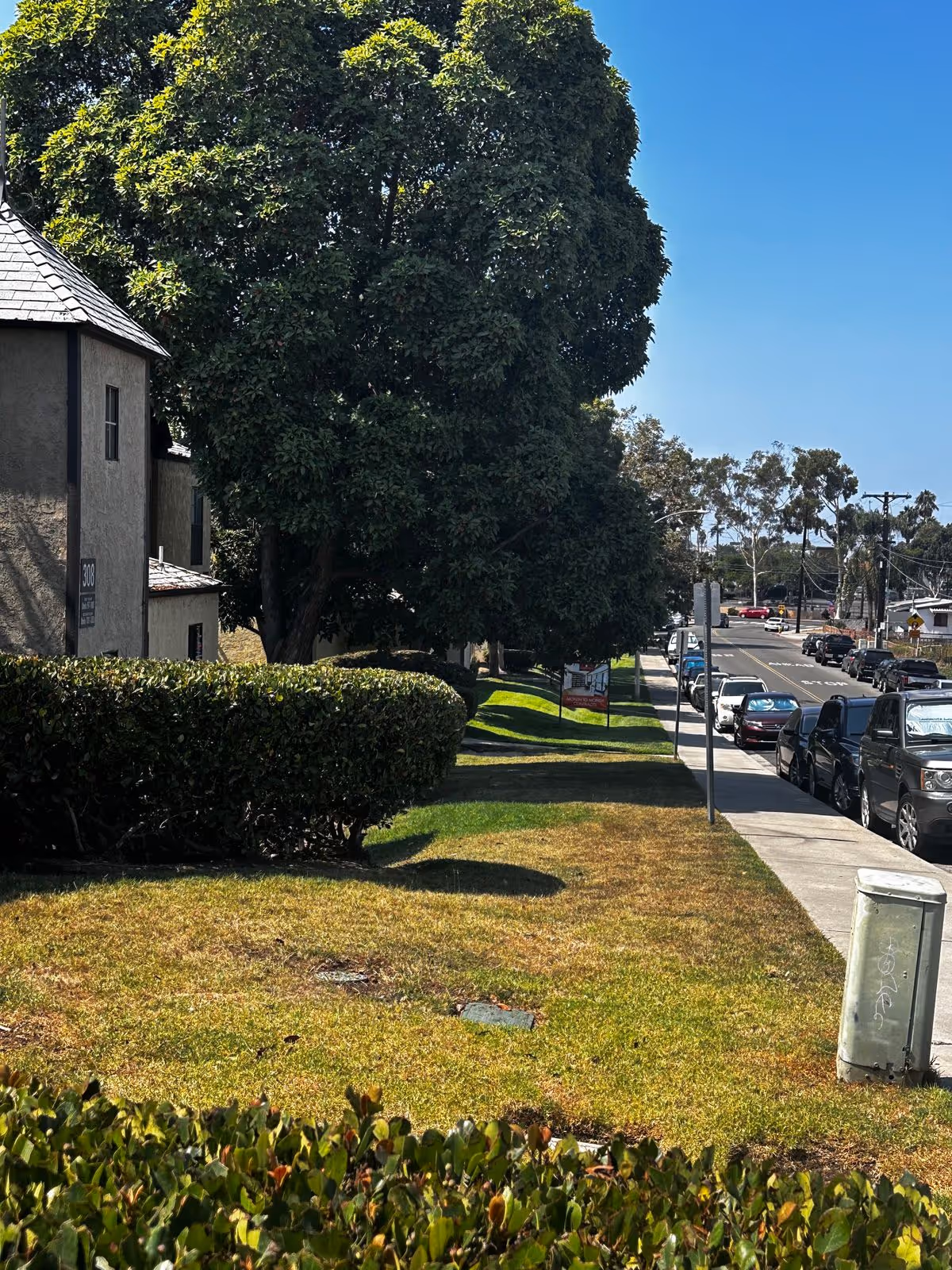 Sidewalk and street beside a two-story apartment building with trimmed hedges, large trees, and parked cars.