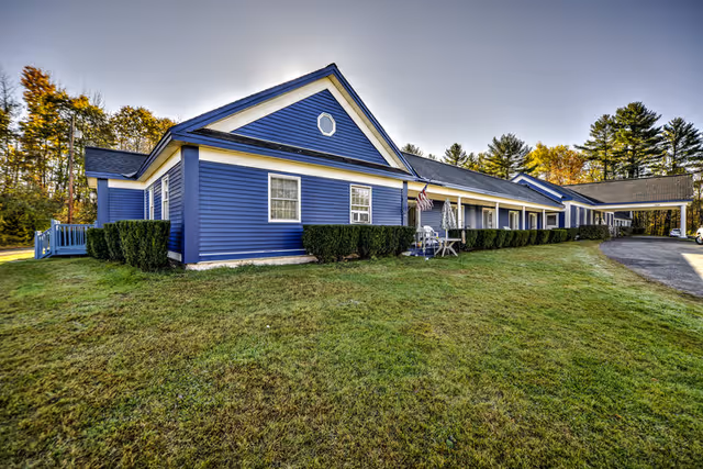 Exterior view of a single-story blue building with white trim, surrounded by green grass and trees in the background under a clear sky. The building has multiple windows, a covered entrance with a driveway, and some outdoor chairs and an umbrella near the entrance.