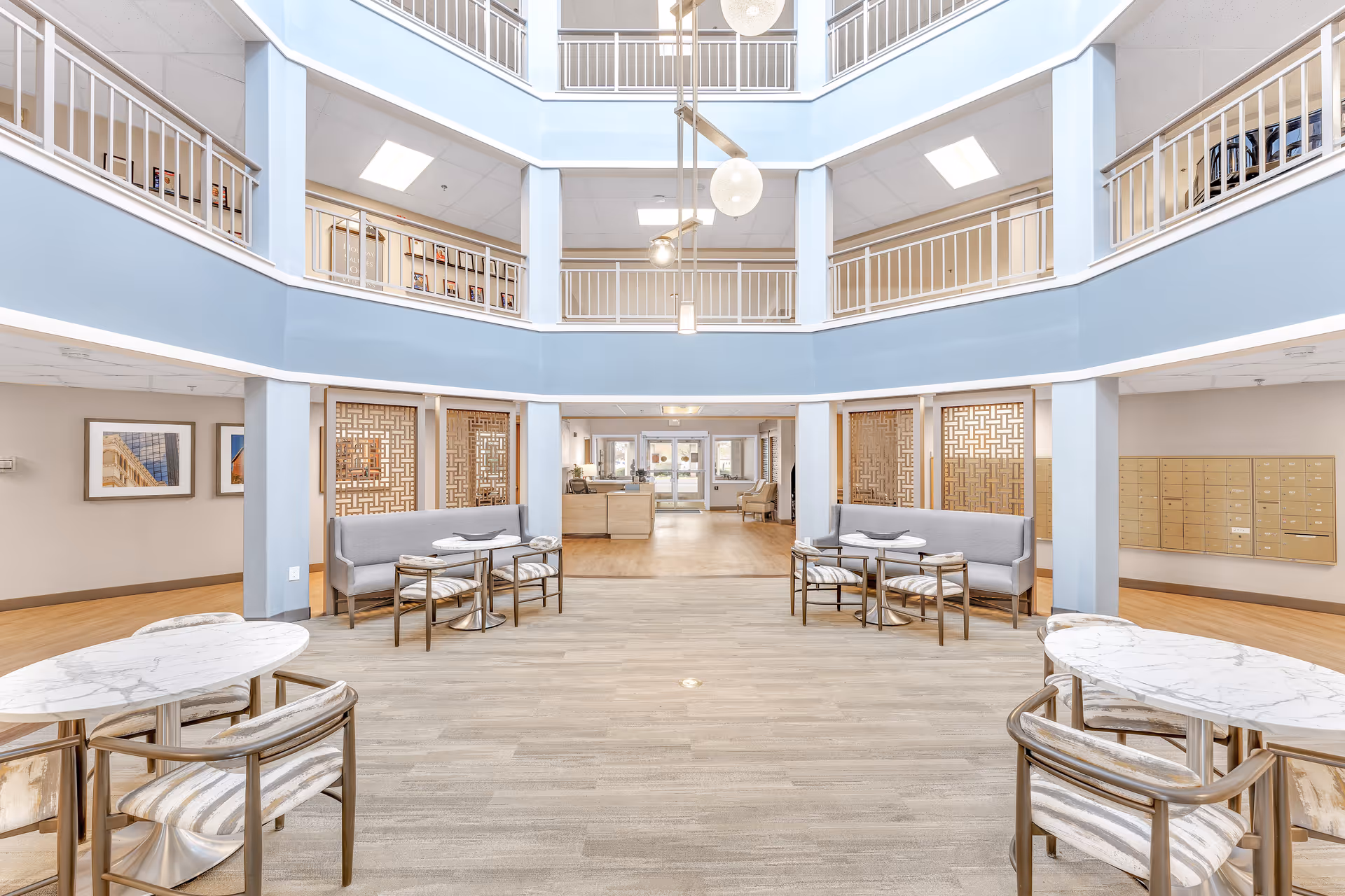 Bright and spacious interior common area of a senior living facility with high ceilings and multiple floors visible. The room features light blue walls, modern pendant lighting, marble-top tables with chairs, and cushioned bench seating. Mailboxes are mounted on the wall to the right, and framed artwork is visible on the left wall.