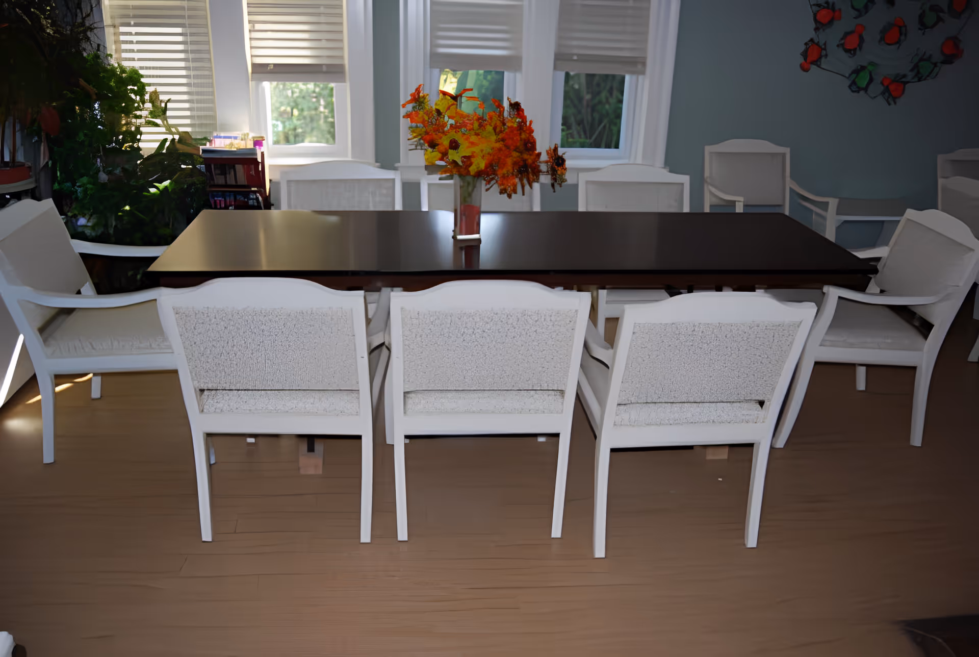 A dark wood dining table surrounded by white upholstered chairs with a vase of autumn flowers in front of windows.