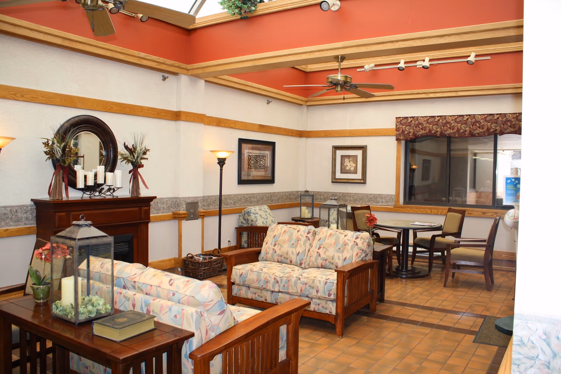 A cozy living room area in Northgate Health Care Facility featuring two floral-patterned sofas with wooden frames, a wooden coffee table with decorative items including a large lantern and a book, a fireplace with a round mirror and candle holders on the mantel, a floor lamp, framed artwork on the walls, ceiling fans, and a small dining table with four chairs near a window.