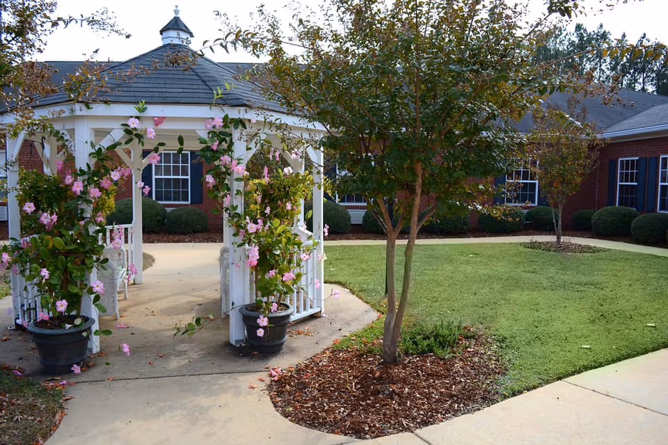 White gazebo covered in pink flowering vines sits in a courtyard with a tree and lawn in front of a red brick building.