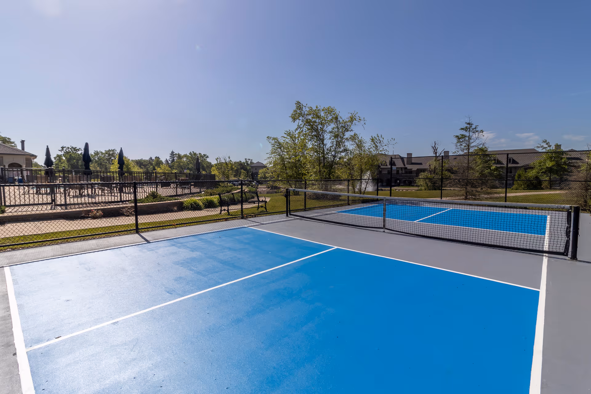 Outdoor view of a blue and gray pickleball court with a net in the middle, surrounded by a black chain-link fence. In the background, there are trees, benches, and buildings under a clear blue sky.