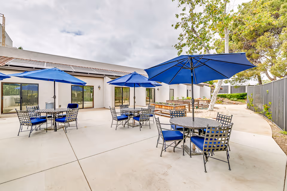 Outdoor patio courtyard with round metal tables, blue umbrellas, and chairs outside a senior living building.