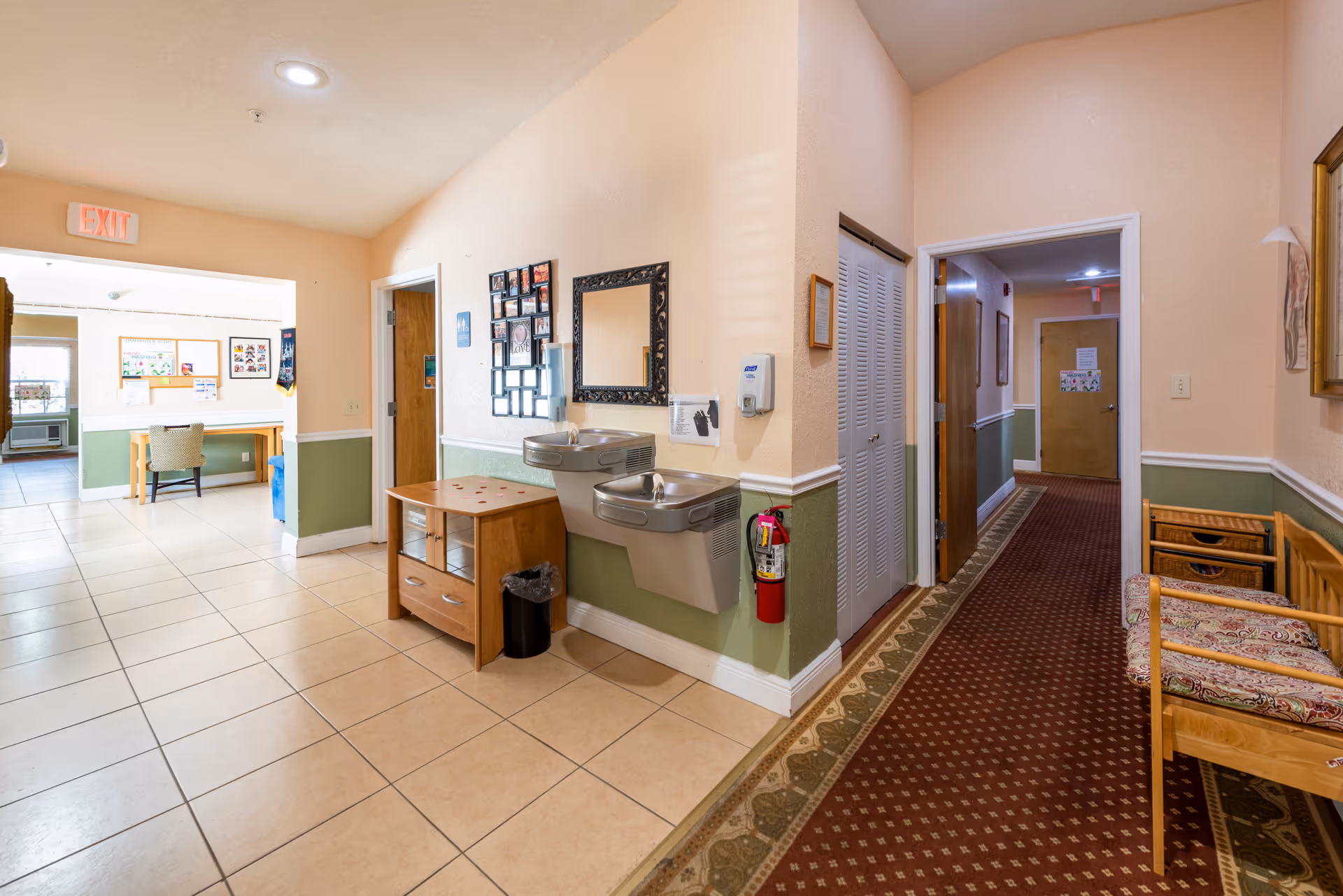 Interior hallway of an assisted living facility with beige and green walls, tiled floor on the left side and carpeted floor on the right side. There are two drinking fountains mounted on the wall, a wooden bench with patterned cushions, a small wooden table with a trash bin underneath, and framed pictures and a mirror on the wall. The hallway leads to other rooms and an exit door is visible in the background.