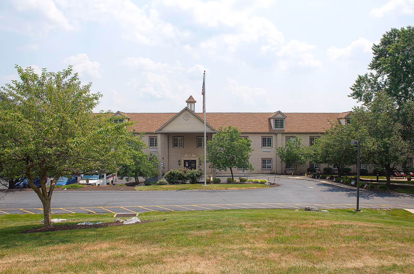 Exterior view of a two-story senior living facility building with a pitched roof, several windows, and a central entrance. The building is surrounded by trees and a grassy area with a paved driveway and parking spaces in front. A flagpole stands near the entrance.