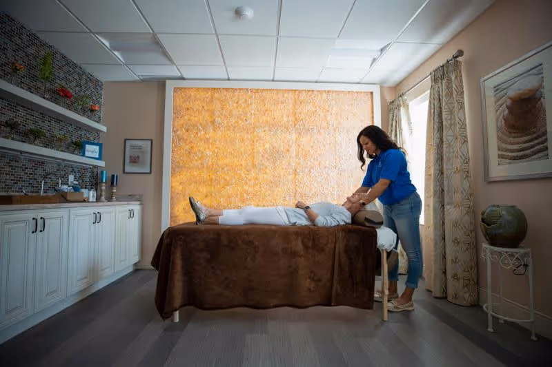 A woman in a blue shirt providing a facial massage to an elderly person lying on a massage table covered with a brown blanket in a softly lit room with decorative curtains, a large textured wall panel, and shelves with small plants and items.