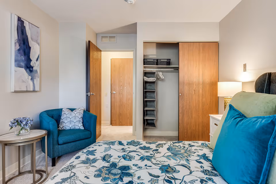 A bedroom in Westhaven Manor featuring a bed with floral bedding and blue pillows, a teal armchair with a patterned cushion, a small round side table with a vase of flowers, an open closet with wooden sliding doors, and a lamp on a white nightstand. A piece of abstract artwork hangs on the wall.