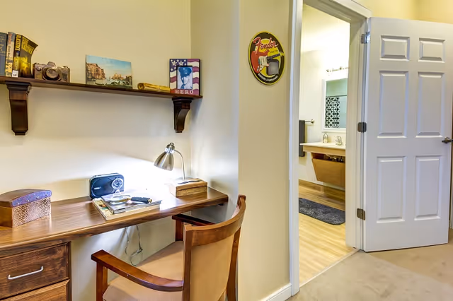 Small workspace with a wooden desk, chair, and lamp beside an open doorway showing a bathroom.