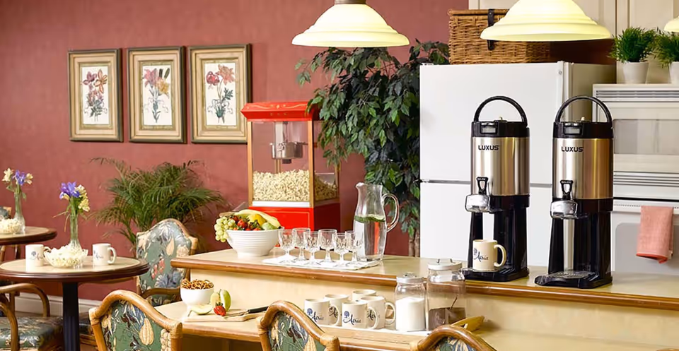 A cozy dining area with round tables and floral upholstered chairs. On the tables are small vases with flowers and mugs. In the background, there is a red popcorn machine, a bowl of fruit, a pitcher of water, and two large coffee dispensers labeled 'LUXUS' on a counter. The walls are decorated with framed floral artwork and there are green plants adding to the ambiance.