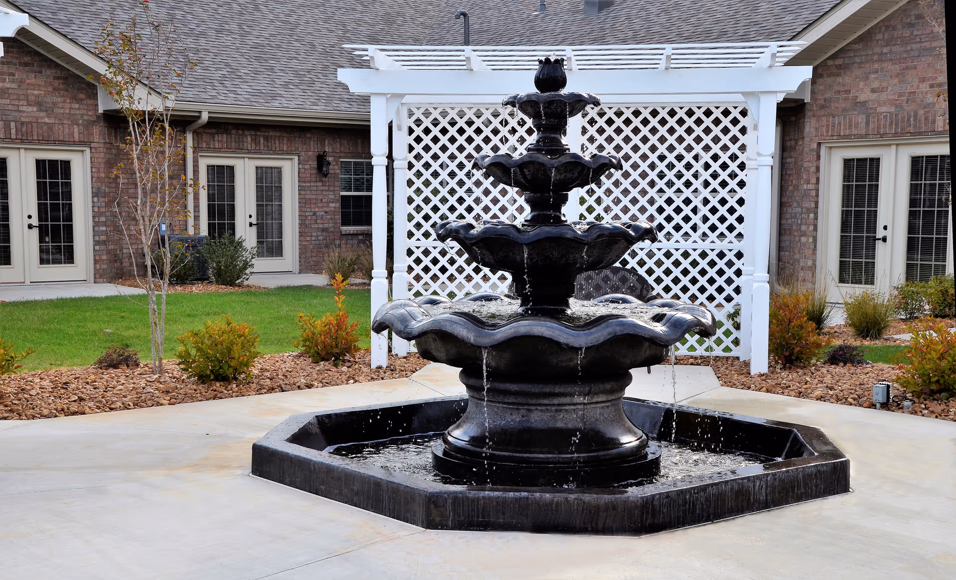 A black tiered water fountain in the center of a paved courtyard area with a white lattice structure and brick building with multiple glass doors in the background. There are small bushes and a young tree planted around the courtyard.