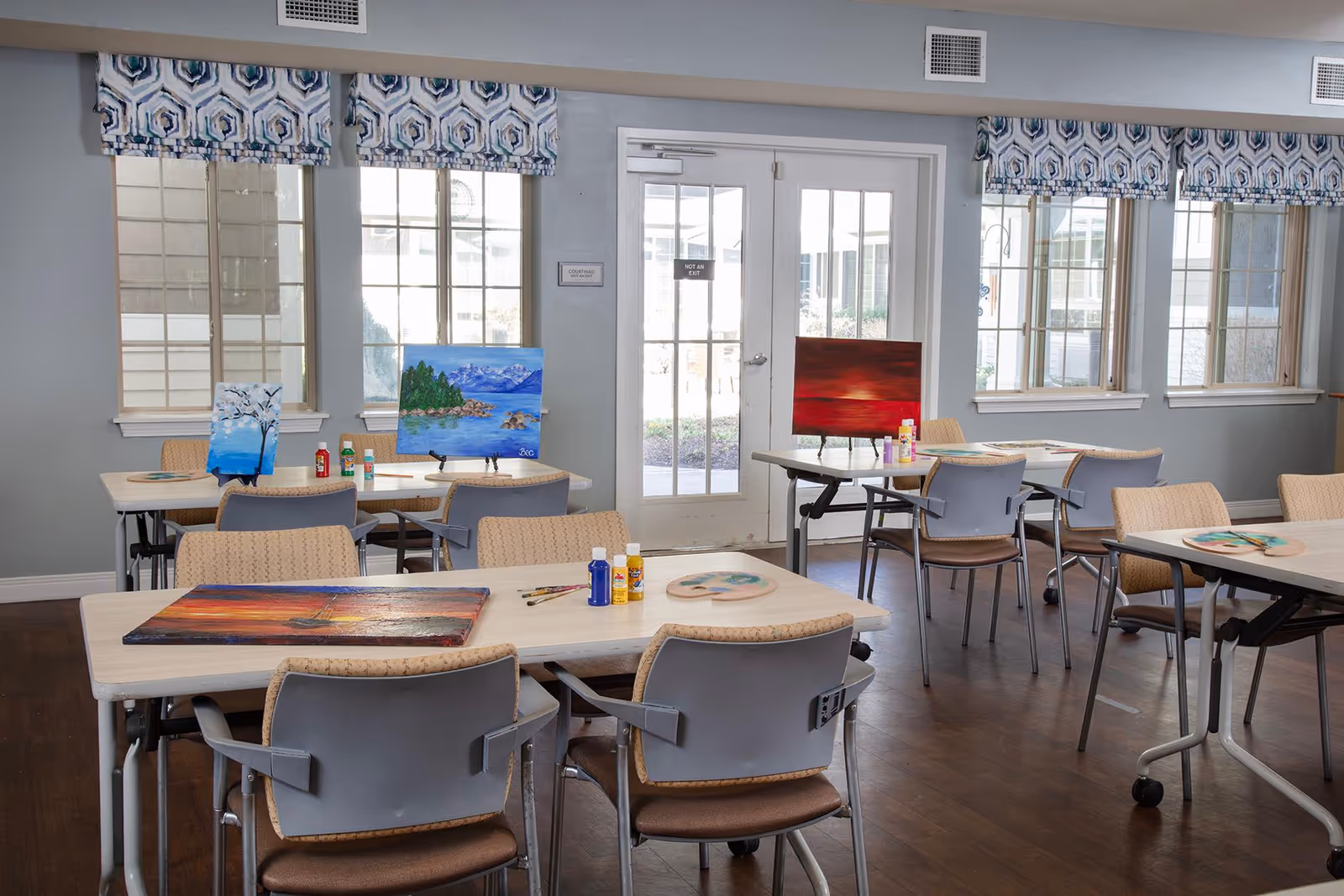 A bright activity room with several tables and chairs arranged for painting. There are colorful paintings on canvases displayed on the tables, along with paint bottles and brushes. The room has large windows with patterned valances and a glass door leading outside.