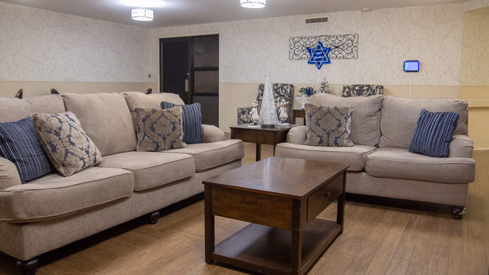 A cozy living room area with two beige sofas adorned with patterned and striped throw pillows. A wooden coffee table with a drawer is placed in front of the sofas. In the background, there is a small table with decorative holiday items including a silver Christmas tree and a menorah. The walls have a light textured wallpaper, and a blue Star of David decoration is mounted on the wall. The floor is wooden, and ceiling lights illuminate the room.