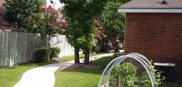 Curved concrete walkway through a grassy courtyard between a brick cottage and a wooden fence with trees and garden beds.