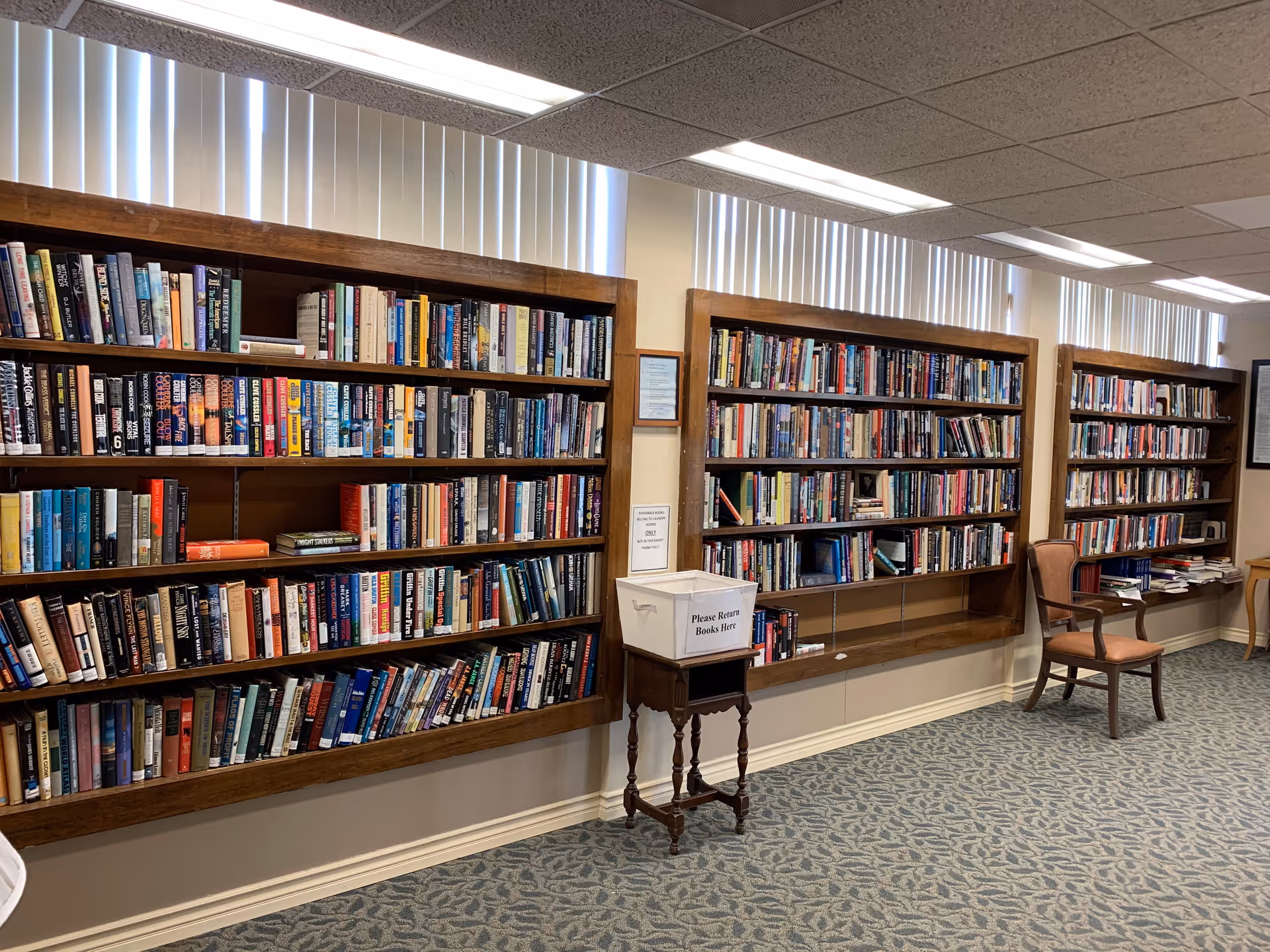 Interior view of a library room with three large wooden bookshelves filled with books mounted on the wall. There is a small wooden table with a white box labeled 'Please Return Books Here' and a wooden chair with a cushioned seat. The room has carpeted flooring and vertical blinds covering the windows near the ceiling.