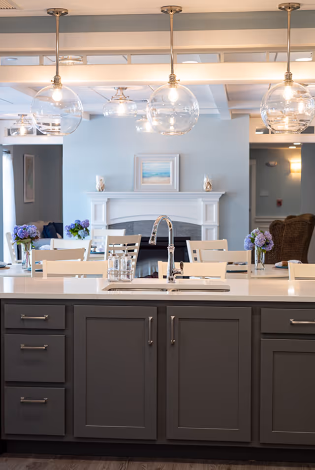 Modern kitchen island with a sink and faucet in the foreground, featuring dark gray cabinets and a white countertop. In the background, there is a dining area with white chairs and tables decorated with purple flowers in vases. A white fireplace with a framed beach painting above it is visible on the light blue wall. The ceiling has multiple round glass pendant lights hanging down.