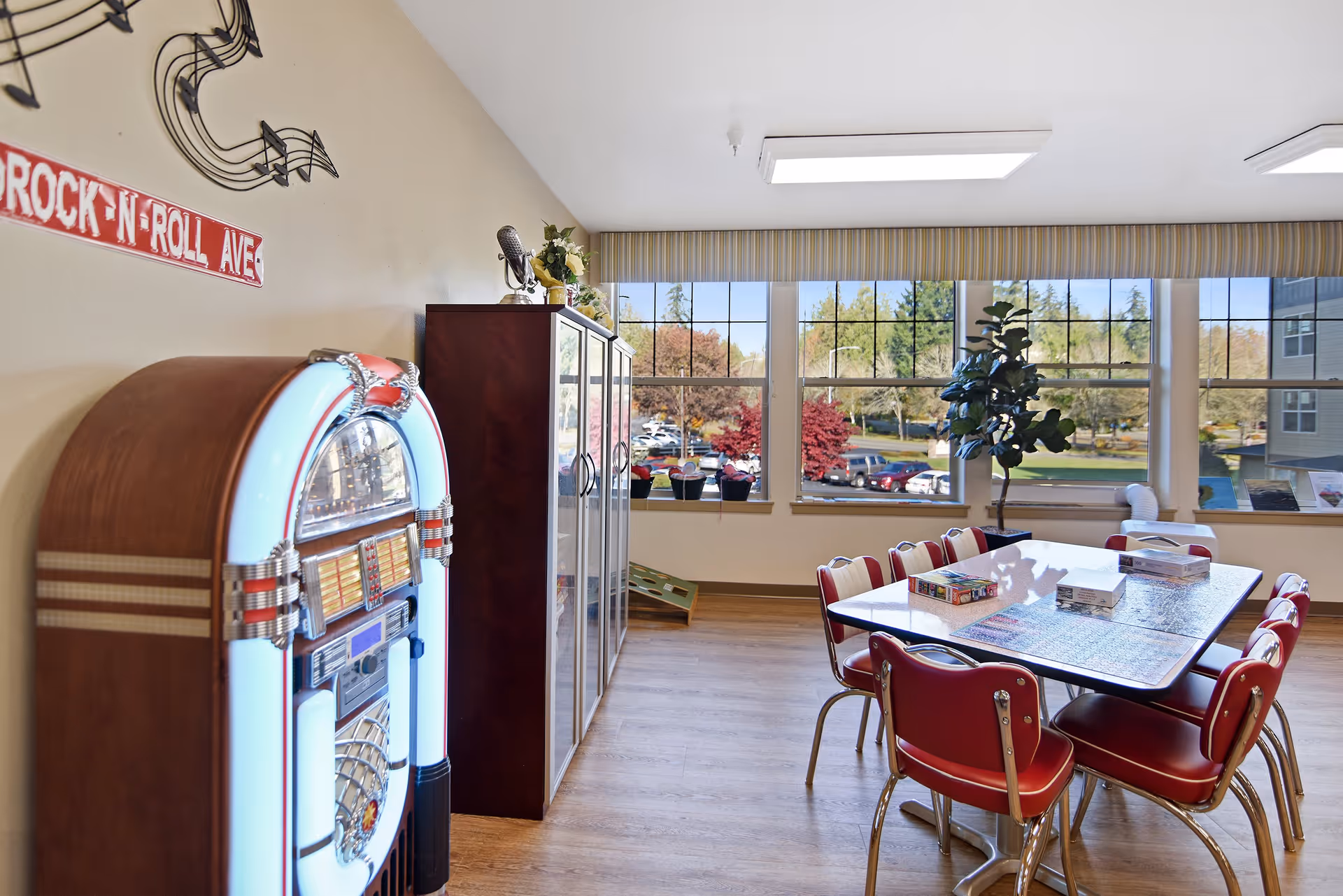 A bright room with large windows showing a view of trees and parked cars outside. Inside, there is a table with red chairs around it, some puzzle boxes on the table, a tall cabinet, a potted plant, and a vintage-style jukebox against the wall. Musical notes and a 'Rock 'N Roll Ave' sign decorate the wall.