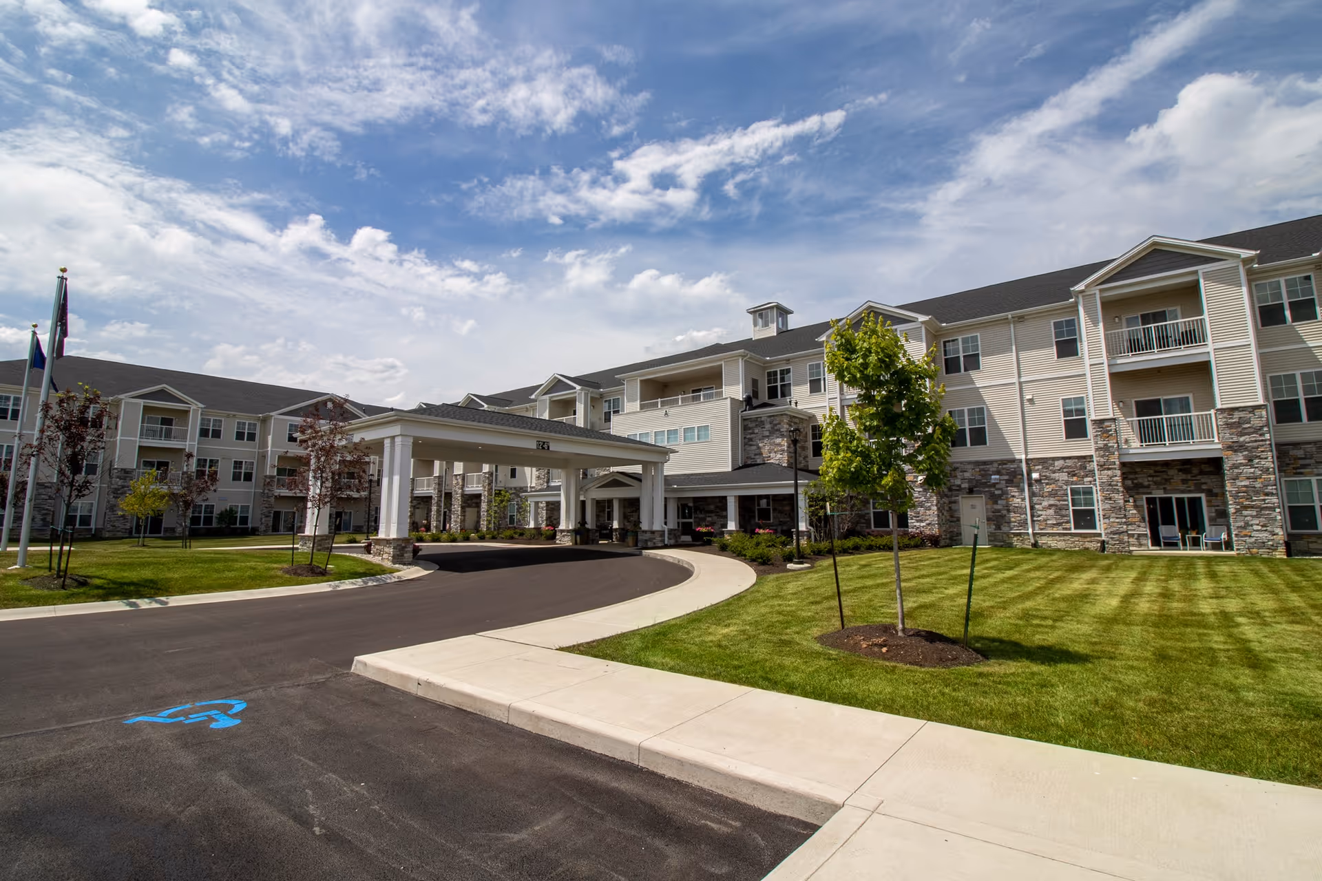 Exterior view of a large, modern senior living facility building with multiple floors, balconies, and a covered entrance driveway. The building is surrounded by well-maintained green lawns, young trees, and a clear blue sky with some clouds.