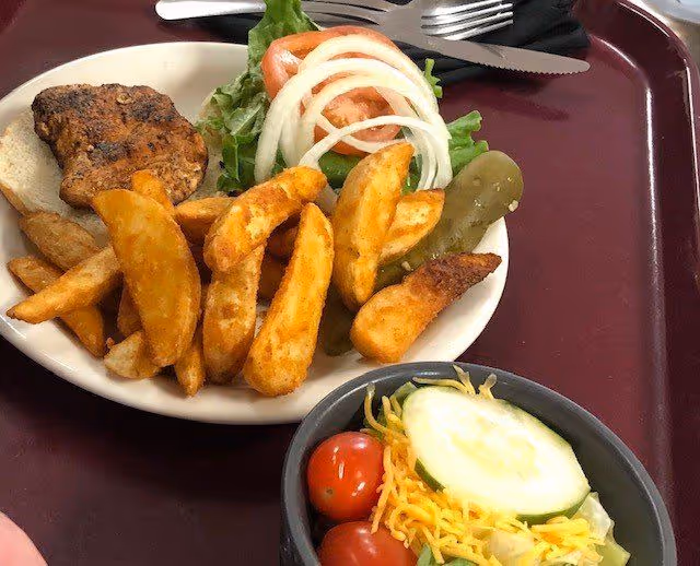 Plate of potato wedges, a small grilled burger patty with lettuce, tomato, onion and a pickle, plus a side salad with cherry tomatoes, cucumber and shredded cheese on a maroon tray.