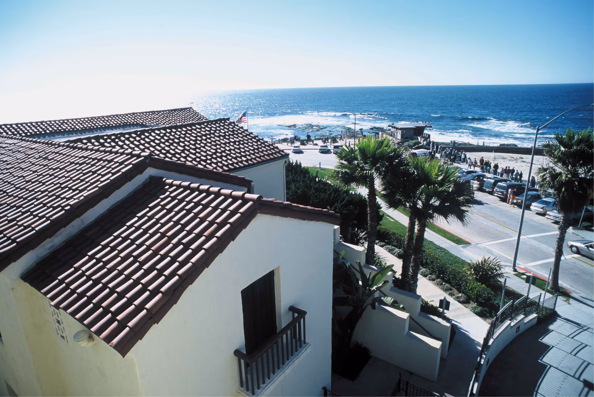 View from above of a building with red tile roofs near a coastal road lined with palm trees, overlooking the ocean with waves and a clear blue sky.