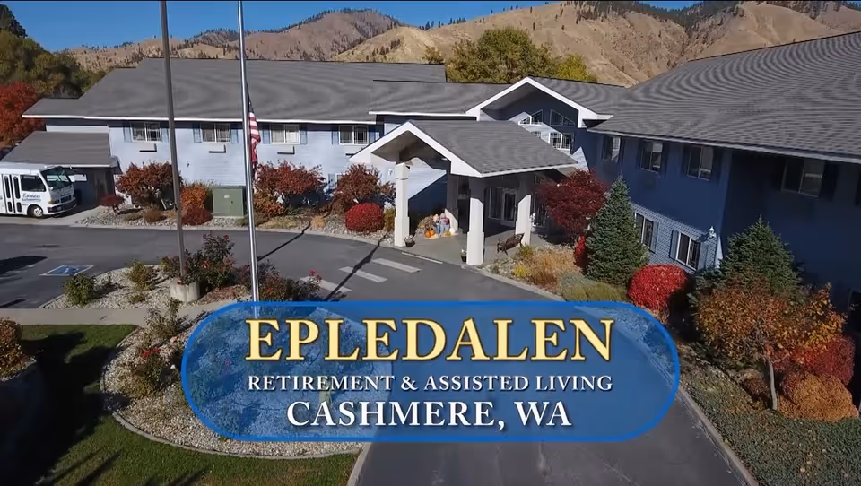 Exterior view of Epledalen Retirement and Assisted Living facility in Cashmere, Washington, showing a two-story building with a covered entrance, landscaped garden with bushes and trees, and a driveway with a parked shuttle bus.