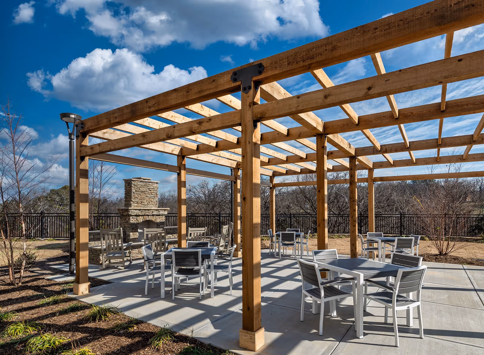 Outdoor seating area with multiple tables and chairs under a wooden pergola. There is a stone fireplace in the background and a black metal fence enclosing the area. The sky is blue with scattered clouds.