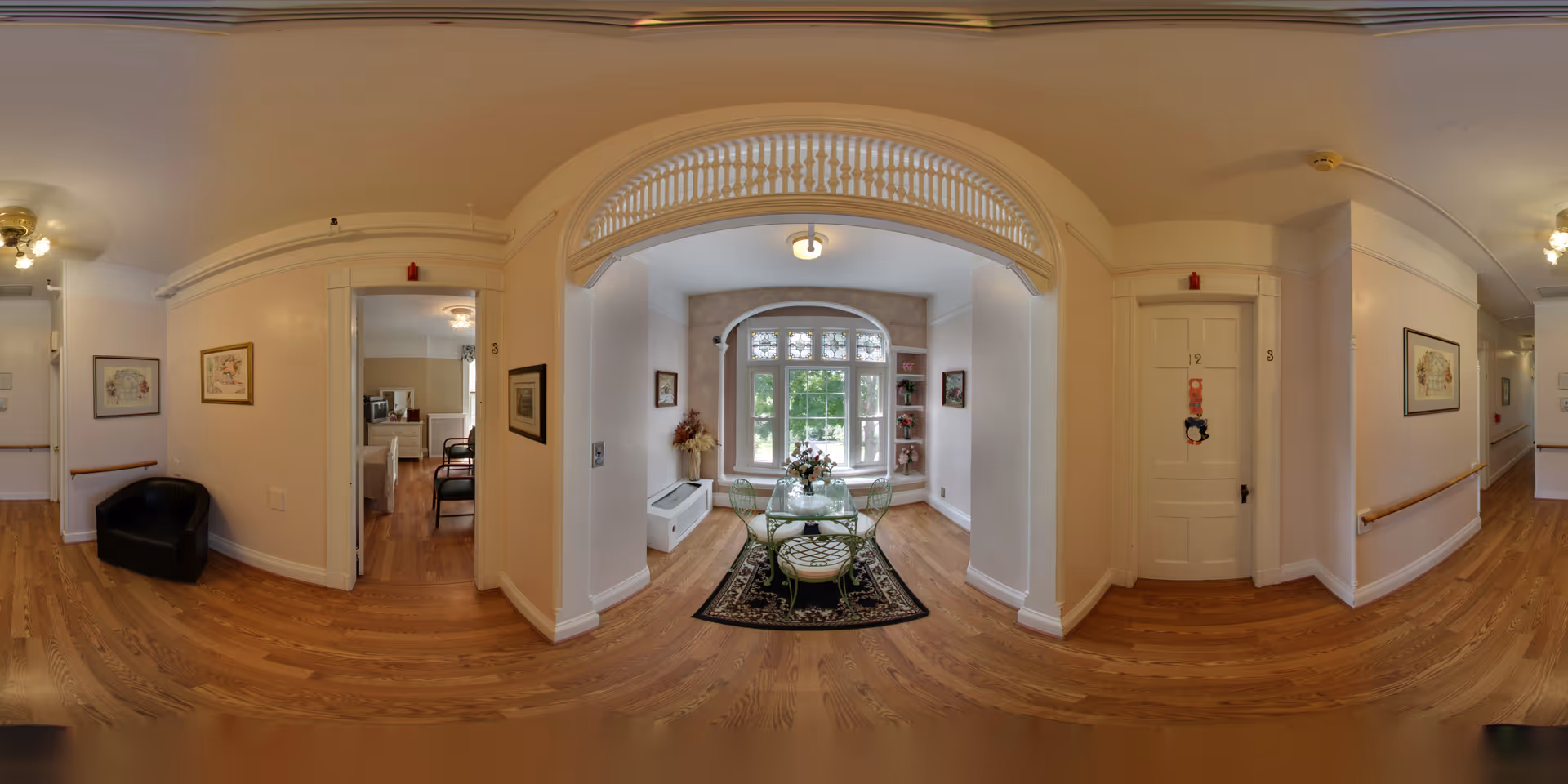 Bright interior hallway with hardwood floors opening onto a small seating area by a large bay window and numbered doors.