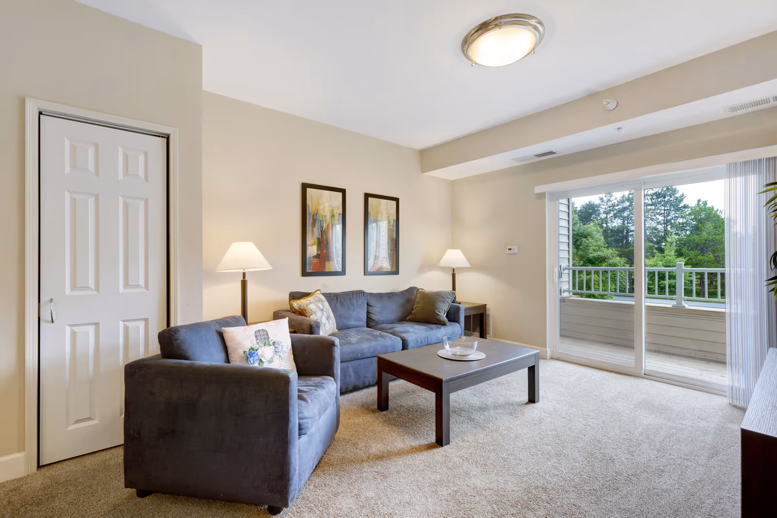 A bright living room with beige walls and carpet, featuring a gray sofa and matching armchair with decorative pillows. There are two framed abstract paintings on the wall above the sofa, two floor lamps on either side of the sofa, a wooden coffee table with a glass bowl, and a sliding glass door leading to a balcony with a view of green trees outside.
