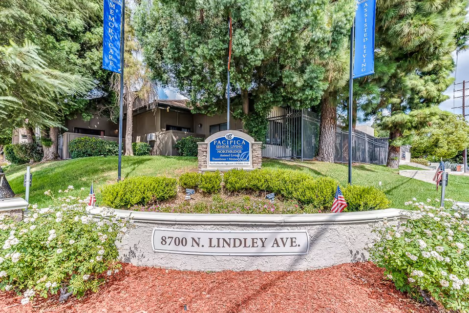 Entrance sign for Pacifica Senior Living Northridge, located at 8700 N. Lindley Ave, surrounded by greenery, bushes, and trees with two blue flags on poles indicating Memory Care and Assisted Living.