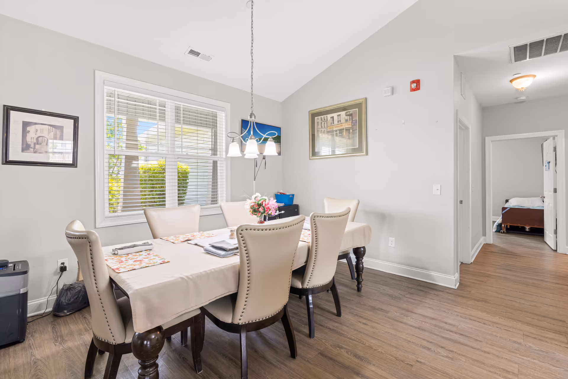 A bright dining room with a rectangular table covered with a beige tablecloth and six cream-colored upholstered chairs. A small floral centerpiece is on the table. The room has light gray walls, a large window with white blinds, two framed pictures on the walls, and a hanging light fixture above the table. A hallway leads to a bedroom with a visible bed in the background.