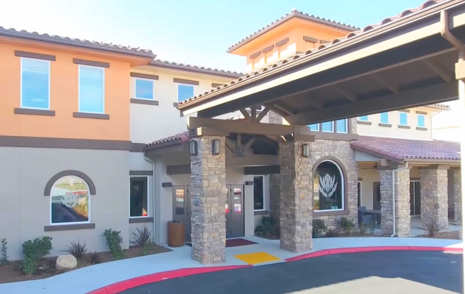 Exterior view of Vineyard Ranch at Temecula senior living facility showing the entrance with stone pillars, a covered driveway, and windows with decorative trim. The building has a mix of beige and orange walls with a tiled roof.