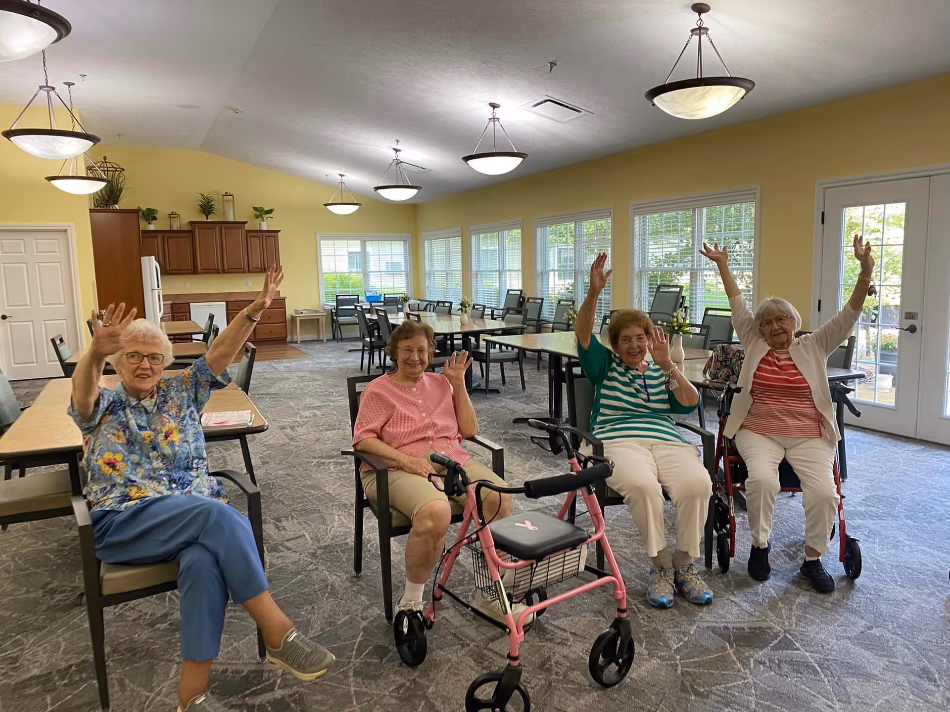 Four women seated and waving in a bright communal dining/activity room with tables, chairs, large windows, and a kitchenette in the background.