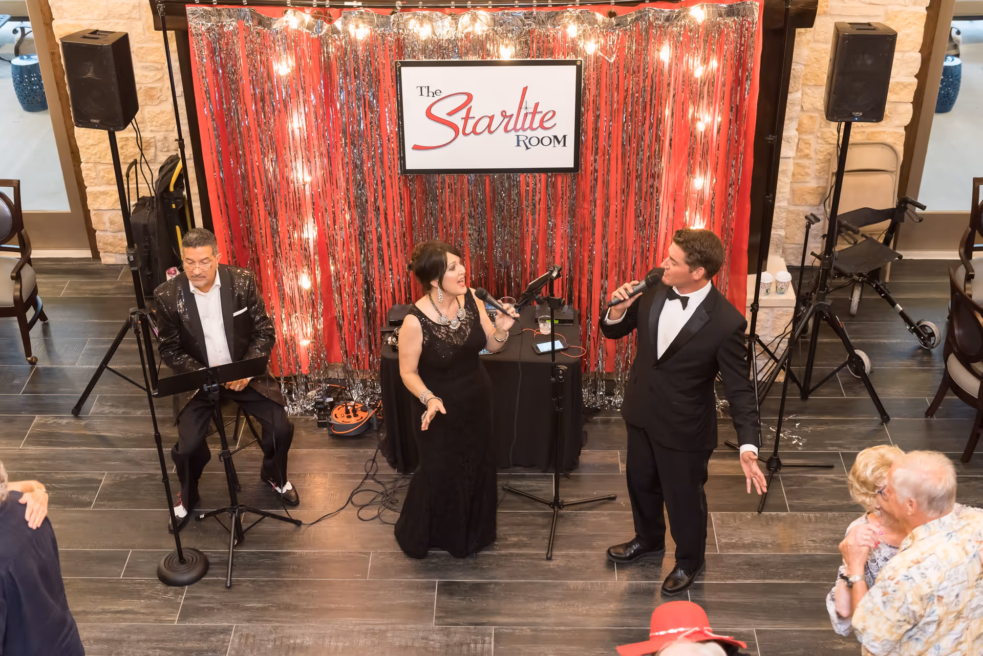 A man and a woman dressed formally are singing into microphones in front of a red and silver tinsel backdrop with a sign that reads 'The Starlite Room.' Another man is seated playing a keyboard. Several elderly people are dancing nearby in a room with stone walls and dark tiled floors.