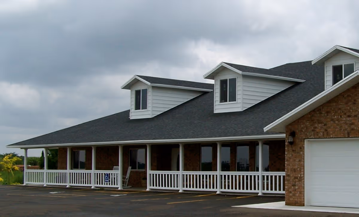Exterior view of a single-story brick building with a dark shingled roof and white trim. The building features a covered porch with white railings and several windows. There are dormer windows on the roof and a garage door on the right side. The sky is cloudy and the parking lot in front is empty.