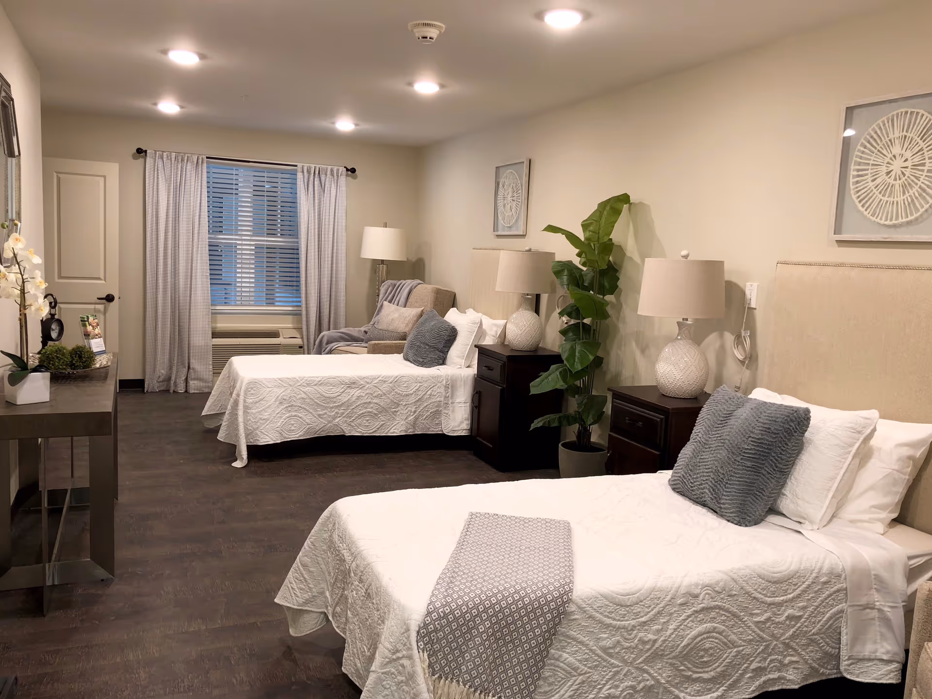 A well-lit bedroom with two twin beds, each with white quilted bedspreads and gray accent pillows. Between the beds are two dark wooden nightstands with matching white lamps. A tall green plant is placed next to one nightstand. At the far end of the room, there is a window with white curtains and an air conditioning unit below it. A beige armchair with a gray throw blanket is positioned near the window. The room has light-colored walls and dark wood flooring.