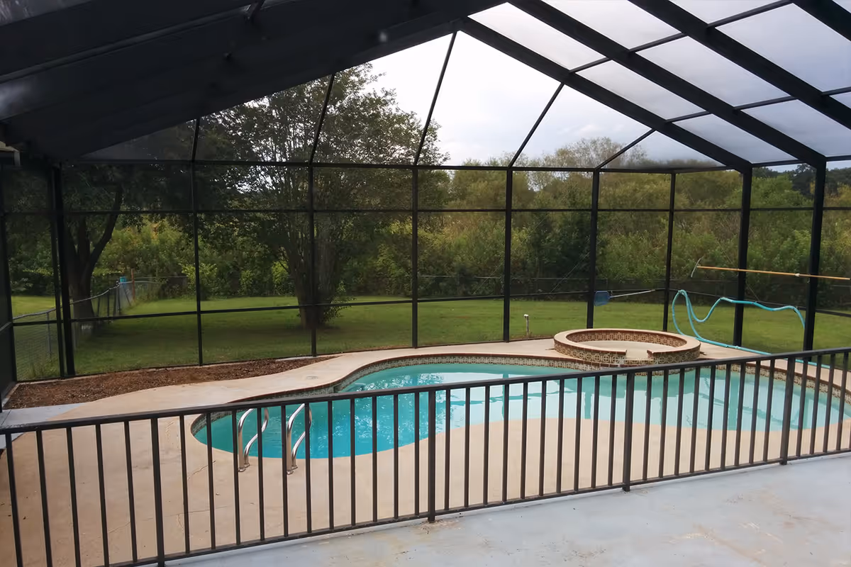 A screened-in outdoor pool area with a kidney-shaped swimming pool and an attached circular hot tub. The pool is surrounded by a concrete deck and enclosed by a black metal fence. Beyond the screened enclosure, there is a grassy yard with trees and shrubs.