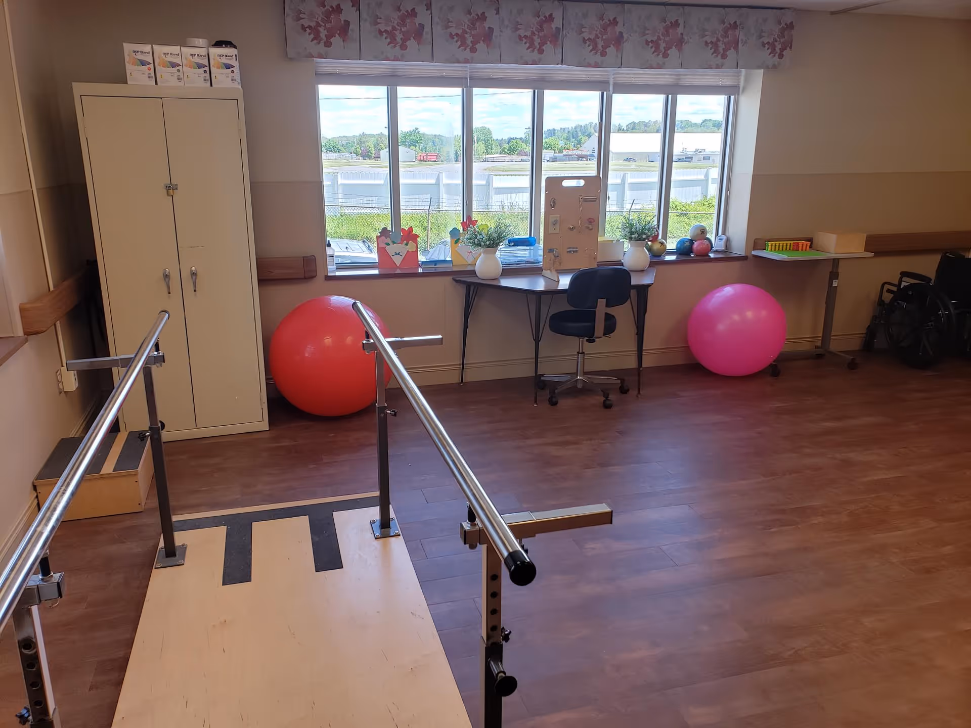 A therapy room with parallel bars for walking exercises, two large exercise balls, a desk with a chair, plants, and therapy tools on the windowsill. There is a cabinet on the left side and a wheelchair on the right side near the wall. The room has wooden flooring and a large window letting in natural light.