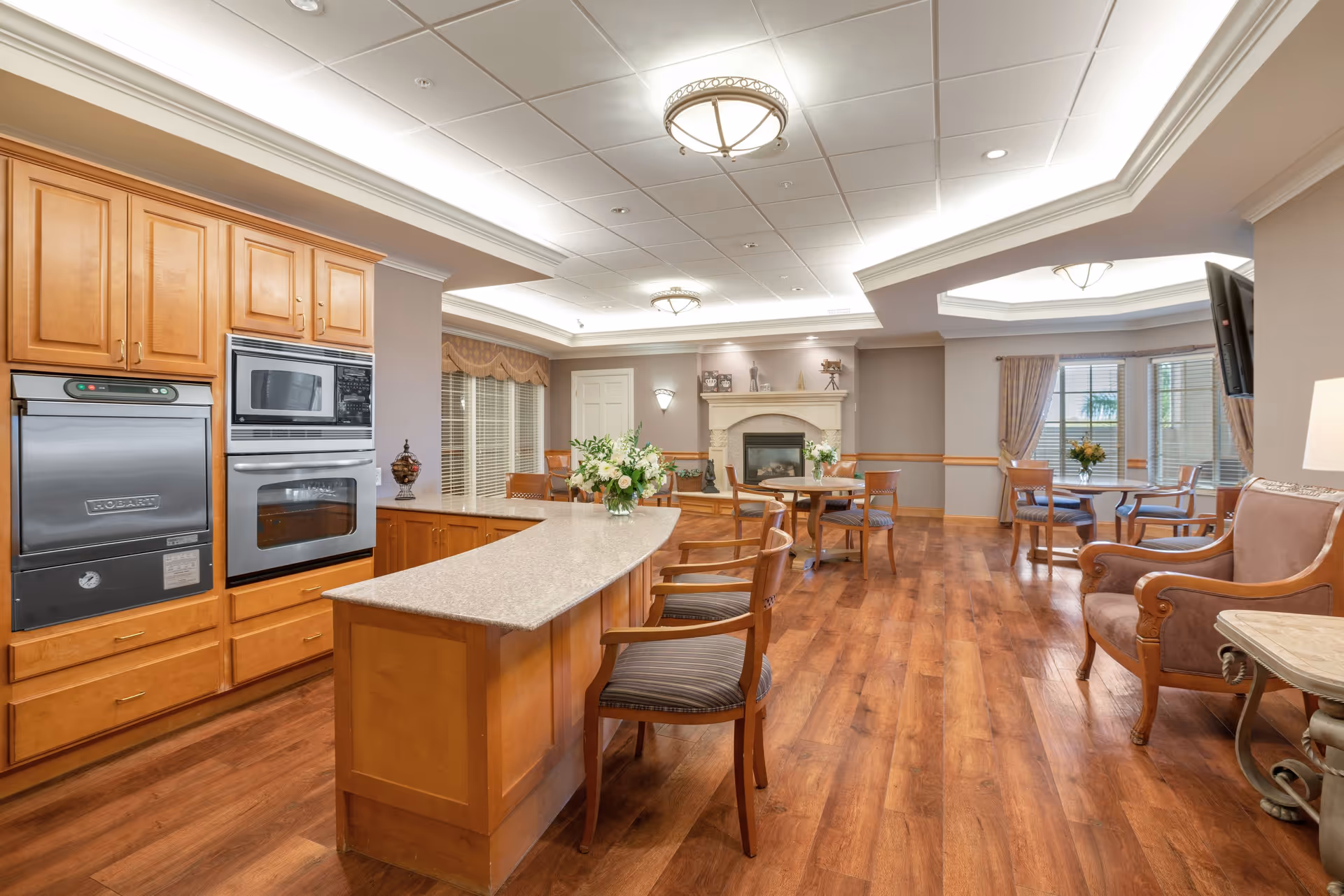 A spacious senior living common area featuring a kitchen with wooden cabinets, built-in oven and microwave, and a long island with chairs. The room has wooden flooring, several round tables with chairs, a fireplace, and large windows with curtains allowing natural light in.