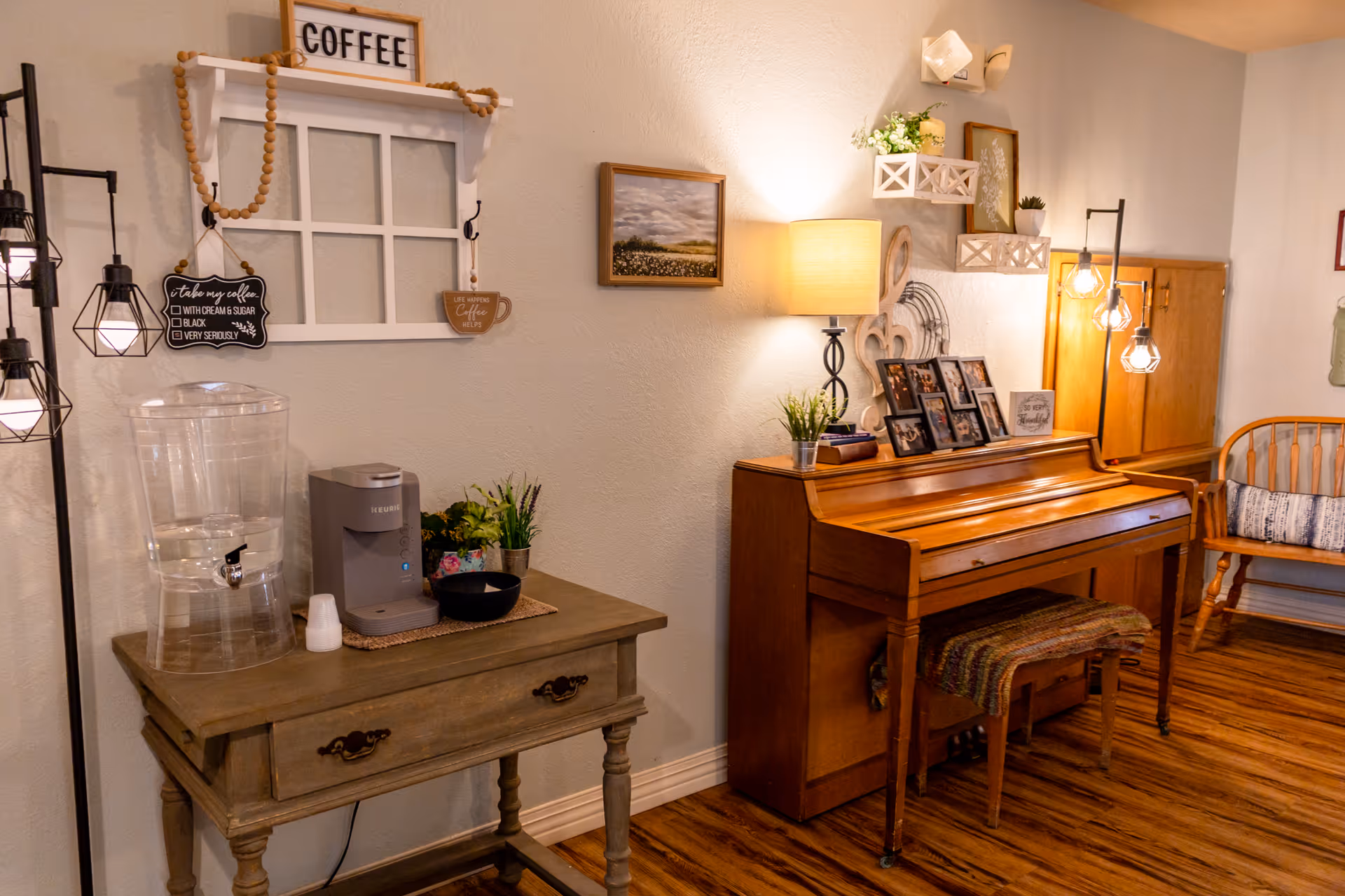 A cozy interior corner of a senior living facility featuring a wooden piano with framed photos and a lamp on top, a rustic wooden table with a water dispenser and coffee maker, decorative wall shelves with plants, and warm hanging lights. The floor is wooden, and there is a wooden bench with a cushion in the background.
