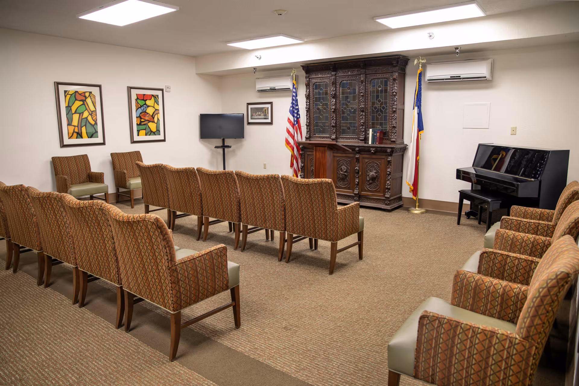 Small meeting room with rows of upholstered chairs facing an ornate wooden cabinet flanked by flags, a TV, and a piano.