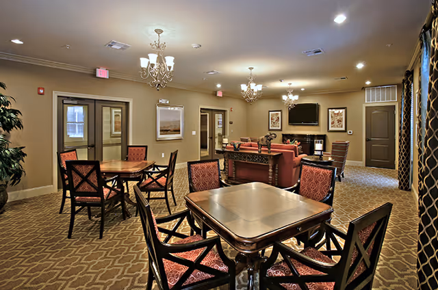 A well-lit common area in a senior living facility featuring multiple wooden tables with red cushioned chairs arranged around them. The room has beige walls, patterned carpet, chandeliers, and a seating area with sofas and a TV mounted on the wall. There are framed pictures on the walls and a large plant in the corner.