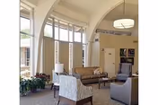 A bright and spacious common area with large arched windows allowing natural light to fill the room. The space features comfortable seating including a beige sofa, patterned armchair, and gray armchair arranged around a coffee table. There are potted plants near the windows and a modern ceiling light fixture hanging from the high ceiling.
